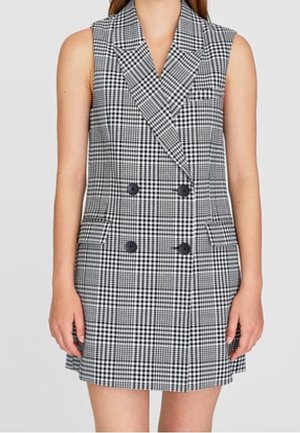 Robe-blazer sans manches à double boutonnage en motif à carreaux, avec de larges revers et deux poches avant, portée par une personne debout.