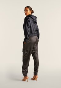 Chaqueta de satén negra con capucha y cintura elástica. Joggers de satén gris oscuro con bolsillos tipo cargo y puños elásticos; combinados con sandalias de tacón color marrón.