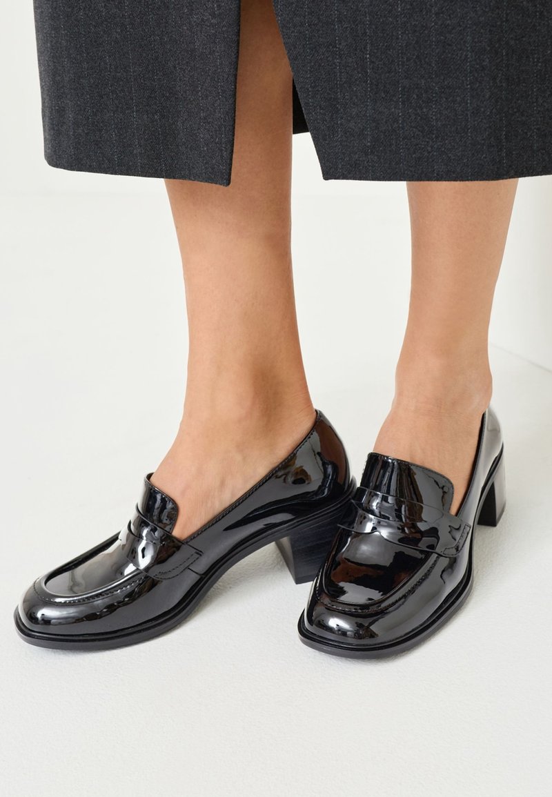 Mocassins à talon bloc en cuir verni noir portés avec un pantalon court gris foncé sur un sol clair.