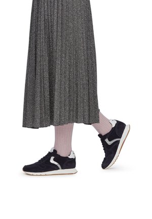 Marineblaue Wildledersneaker mit silbernen Akzenten, weißer Sohle, kombiniert mit hellrosa Socken und einem metallisch plissierten grauen Rock.