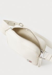 Kremfarget lær crossbody-veske med sølvglidelås, justerbar stropp og logo-patch. Innvendig er den glatt og uten pynt.