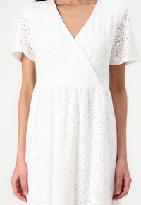 White dress made of eyelet fabric with a wrap design, short sleeves, v-neck, and subtle patterns of dots and swirls.