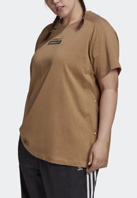 Vrouw in een bruine Adidas t-shirt met logo patch op de borst en zwarte Adidas trainingsbroek met witte strepen aan de zijkanten.