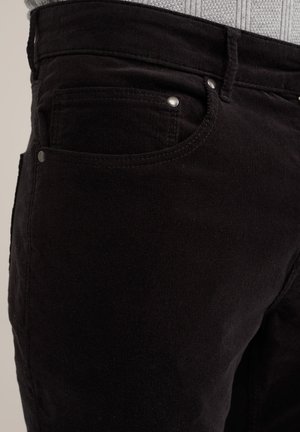 Sorte fløjl bukser med synlig frontlomme, metalhardware accenter og tekstureret stof. Nærbillede, der fremhæver materialet og designet.