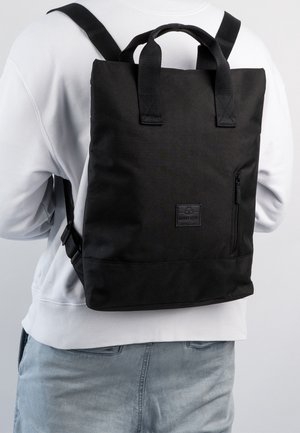 Person iført sort firkantet rygsæk med top-håndtag og en lomme med lynlås foran, iført hvid sweatshirt og lysegrå jeans.