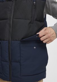 Navy puffer vest with a black, quilted panel, featuring a zippered chest pocket and a button-secured lower pocket. Soft texture.