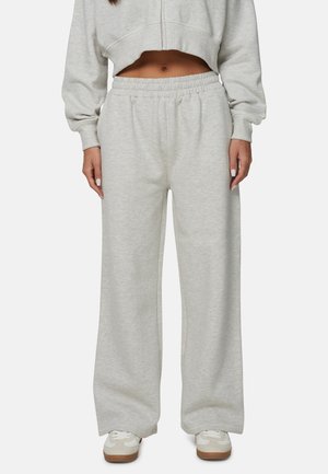 Osoba ubrana w jasnoszare, szerokie spodnie dresowe, krótki kaptur i białe trampki, stojąca na tle jednolitego białego tła.