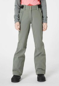 Grüne Softshell-Hose mit glatter Textur, ausgestattet mit Reißverschlusstaschen, einer hohen Taille und bronzenen Knöpfen zum Verschließen. Kombiniert mit schwarzen Stiefeln.