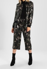 Zwarte bomberjack met pailletten en camouflagepatroon, gecombineerd met bijpassende cropped broeken. Model draagt zwarte enkellaarsjes en toont de gestructureerde stof.