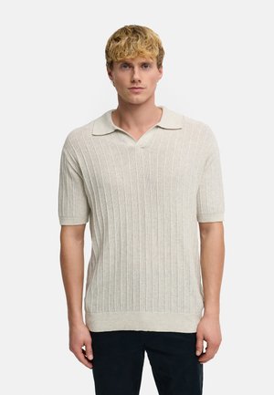 Blonder Mann, der ein kurzärmeliges, cremefarbenes, geripptes Strick-Poloshirt und dunkle Hosen trägt, steht vor einem einfarbigen Hintergrund.