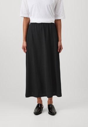 Persoon die een eenvoudig wit shirt met korte mouwen, een zwarte rok tot net boven de enkel en zwarte instapschoenen draagt, staand tegen een neutrale achtergrond.