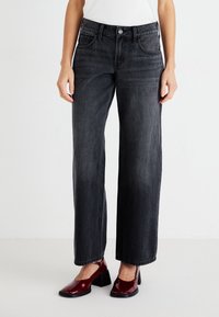 Jeans de mezclilla negros de pierna ancha con cintura alta, diseño de cinco bolsillos y sutil textura desgastada. Combinados con tacones burdeos brillantes.