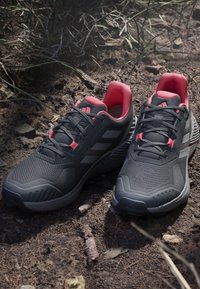 Scarpe sportive nere con accenti rossi, parte superiore in rete testurizzata, suola in gomma spessa e strisce laterali prominenti, posizionate su una superficie naturale.