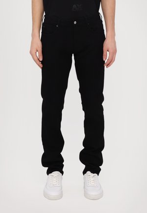 Osoba w obcisłych czarnych dżinsach i białych trampkach, stojąca na tle jednolitego jasnego tła.