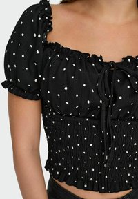 Sort cropped bluse med puffede ærmer, hvide polka dots, smocket tekstur og en snor i halsen med frynsede detaljer.