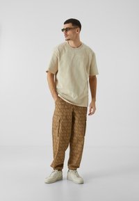 Tricou oversized bej cu mâneci scurte și un logo discret, purtat cu pantaloni cargo maro cu un model de text repetat și adidași albi.