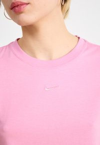 Rožinė medvilninė marškinėliai su apvalia iškirpte, trumpomis rankovėmis ir mažuoju siuvinėtu Nike logotipu, kuris šiek tiek tamsesnis už rožinę.
