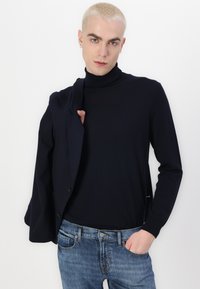 Navy turtleneck sweater in a soft, smooth fabric, paired with a dark blazer draped over the left arm, and light blue jeans with a classic fit.