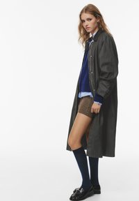 Cappotto lungo, verde scuro con bottoni argentati, indossato sopra un maglione blu e pantaloni corti marroni, abbinato a calze navy fino al ginocchio e mocassini neri.