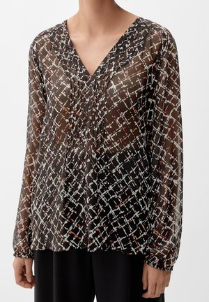 Femme portant un chemisier noir transparent avec un motif quadrillé abstrait blanc et marron et des manches longues.