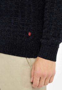 Maglione lavorato a maglia blu scuro con una trama testurizzata, polsini a coste e una piccola patch rossa con logo sull'orlo. Abbinali a pantaloni beige chiaro.