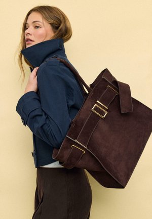 Jeune femme portant une veste bleu marine à col montant et un grand sac cabas en daim brun foncé avec des boucles dorées sur l'épaule.