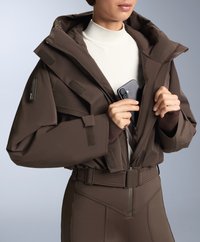 Hnedá zateplená bunda s veľkou kapucňou, so zipsom vpredu a velkro detailmi; nosená na svetlom farebnom roláku, s telefónom v ruke.