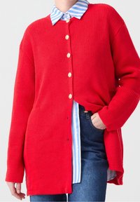 Red ribbed cardigan with large buttons, featuring wide sleeves and a longer cut, layered over a light blue and white striped shirt.