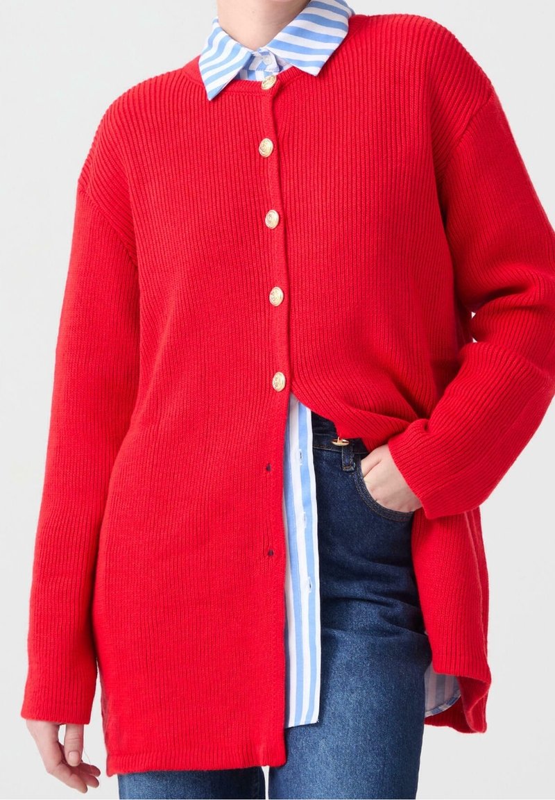 Red ribbed cardigan with large buttons, featuring wide sleeves and a longer cut, layered over a light blue and white striped shirt.