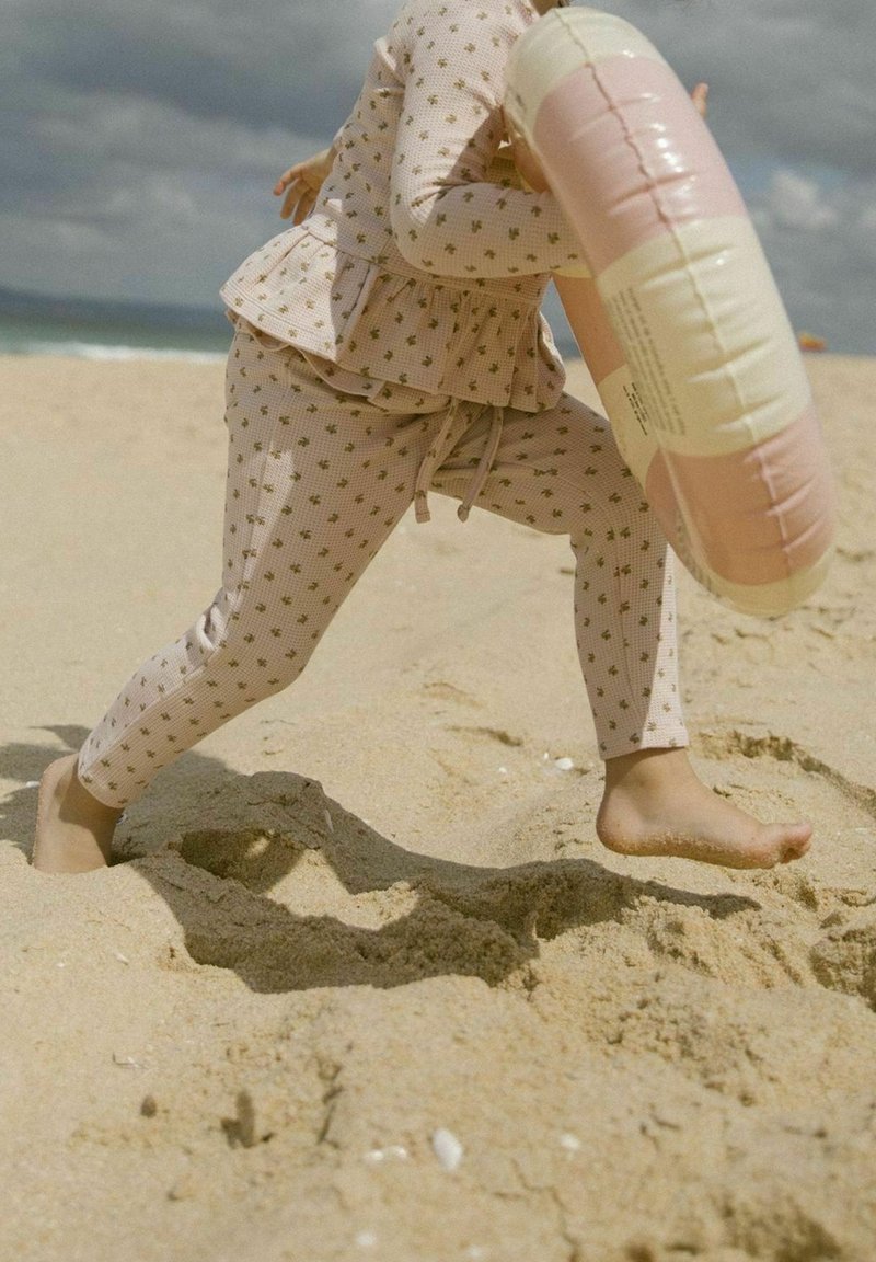 Enfant portant un pyjama à motifs courant pieds nus sur une plage de sable, tenant une bouée gonflable rose et blanche.