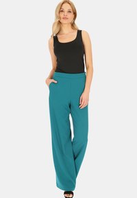 Czarny top na ramiączkach w połączeniu z szerokimi, tealowymi spodniami z bocznymi kieszeniami i gładką teksturą. Model stoi na jednolitym tle.