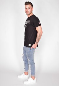 Svart kortärmad t-shirt med "TOP GUN" tryck, parat med ljusblå rivna skinny jeans och vita sneakers, placerad mot en enfärgad bakgrund.