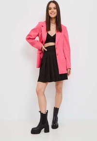 Blazer rosa oversized, top corto negro, falda plisada negra y botas de tobillo de tacón grueso negro. Texturas suaves con detalles de diseño a medida.
