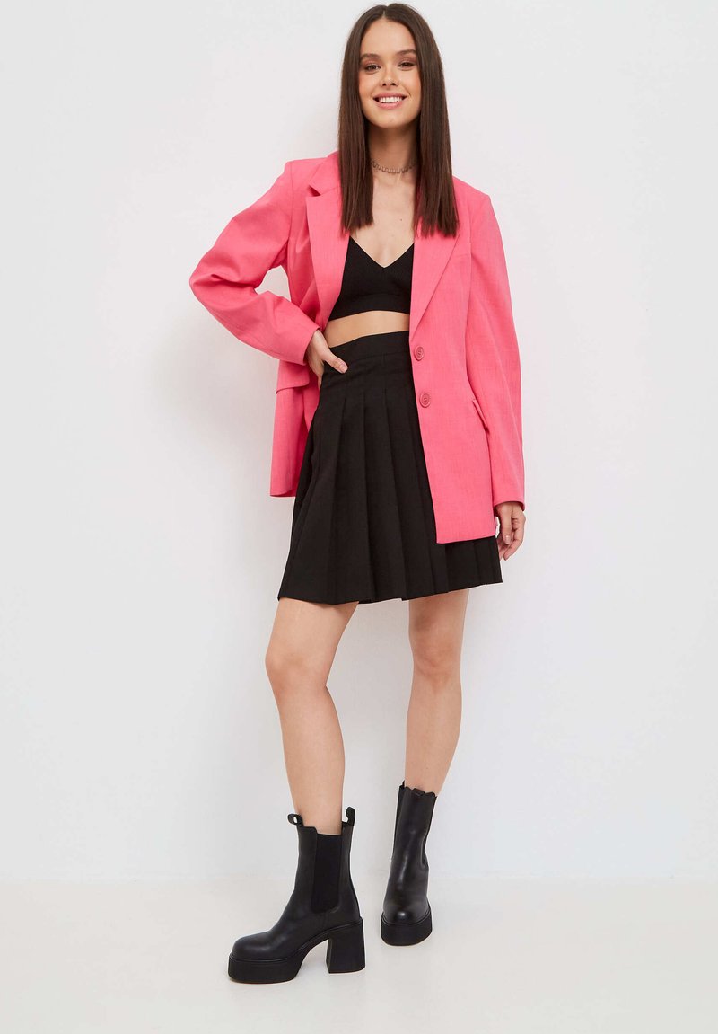 Blazer rosa oversized, top corto negro, falda plisada negra y botas de tobillo de tacón grueso negro. Texturas suaves con detalles de diseño a medida.