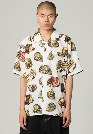 Joven con una camisa blanca de manga corta con estampado de anillos dorados, rostros y piedras preciosas rojas, de pie frente a un fondo liso.