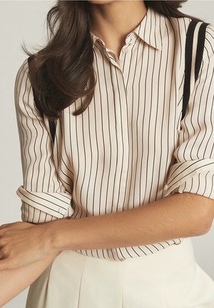 Lichtbeige blouse met verticale zwarte strepen, knoopsluiting en opgerolde mouwen met donkere accenten op de schouders.