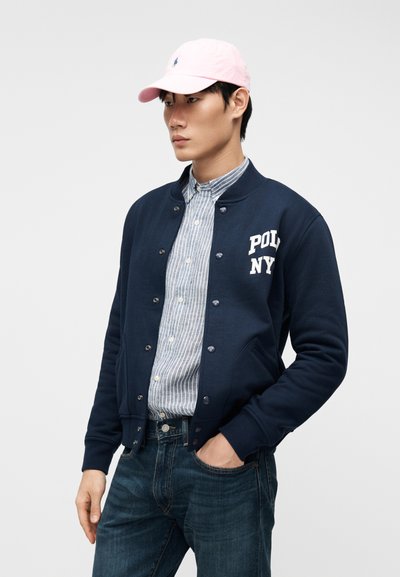 Νεαρός άνδρας με ροζ καπέλο, μπλε σκούρο varsity μπουφάν με κουμπιά και το κείμενο "POL NY", ριγέ πουκάμισο με κουμπιά και μπλε τζιν, με το ένα χέρι στην τσέπη.