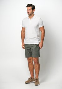 Vit pikétröja med korta ärmar, matchad med gröna shorts. Modellen står i avslappnade skor, med en neutral bakgrund.