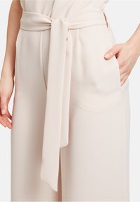 Lys rosa jumpsuit med bælte og struktureret overflade, der har sidelommer og et bånd om taljen for et figursyet udseende.