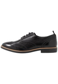Chaussure oxford en cuir noir avec un bout arrondi, des détails de brogue décoratifs et un talon bas empilé. Elle présente une texture douce et des lacets noirs.