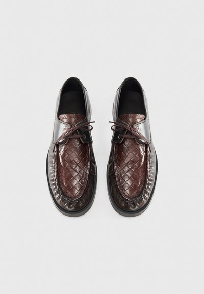 Scarpe in pelle intrecciata marroni con punta lucida marrone scuro, lacci rotondi e forma arrotondata. Accenti neri lisci nella parte del colletto.
