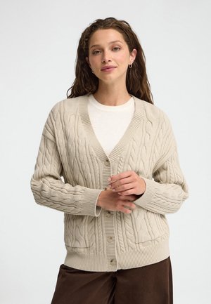 Mujer con el cabello largo y rizado que lleva un cárdigan de punto de cable en crema sobre una camiseta blanca, de pie con las manos suavemente entrelazadas frente a un fondo liso.