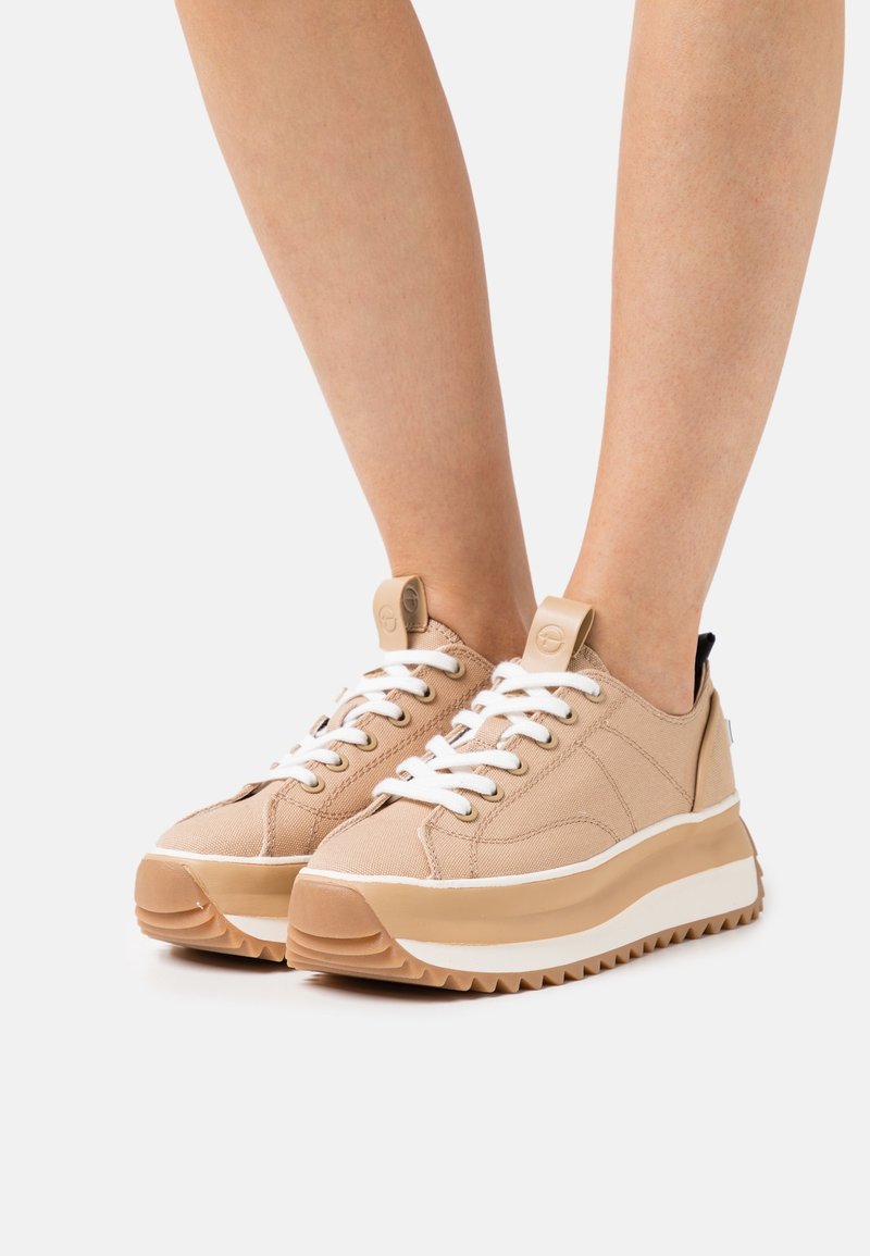 Jambes portant des baskets à plateforme beige avec des lacets blancs et des semelles épaisses, debout sur un fond gris clair.