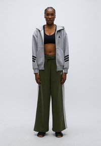 Szara bluza z kapturem na zamek błyskawiczny z trzema czarnymi paskami na rękawach, zestawiona z luźnymi oliwkowozielonymi spodniami dresowymi z białymi bocznymi paskami. Widoczny czarny sportowy stanik.