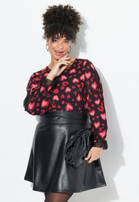 Blusa negra de manga larga con estampados de corazones rojos, combinada con una falda corta de cuero negra con pliegues. El accesorio incluye un bolso de mano negro texturizado.