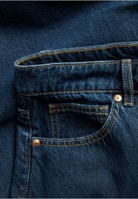 Close-up of blue denim jeans showing front pocket, coin pocket, belt loop, and copper rivets with visible orange stitching.