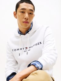 White hoodie featuring "TOMMY HILFIGER" text and logo, with drawstrings. Worn over a blue plaid shirt, with bare forearm visible.