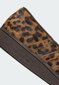 Παπούτσι slip-on με λεοπάρ σχέδιο, επάνω μέρος από υφή γούνας, καφέ λαστιχένια σόλα και λεπτομέρεια λογότυπου στο πλάι. Λεία επιφάνεια με έντονο μοτίβο.