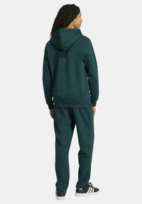 Set da jogging verde scuro in tessuto morbido, composto da felpa con cappuccio e pantaloni abbinati, con polsini a coste e eleganti sneaker nere.