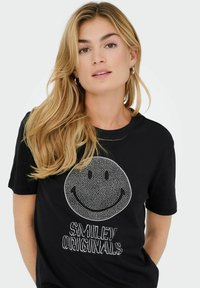 T-shirt en coton noir avec un graphique de visage souriant à motifs de pois blancs et le texte "SMILEY ORIGINALS" dans une police texturée.